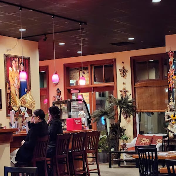 Two people seated at a bar in a Thai restaurant with traditional decor and pink pendant lights.