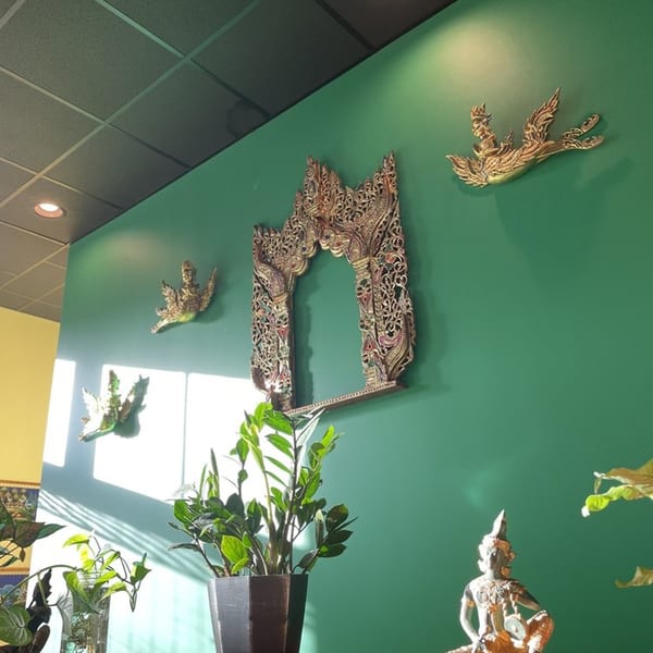 Green wall with ornate golden Thai decor and potted plants in a sunlit restaurant interior.
