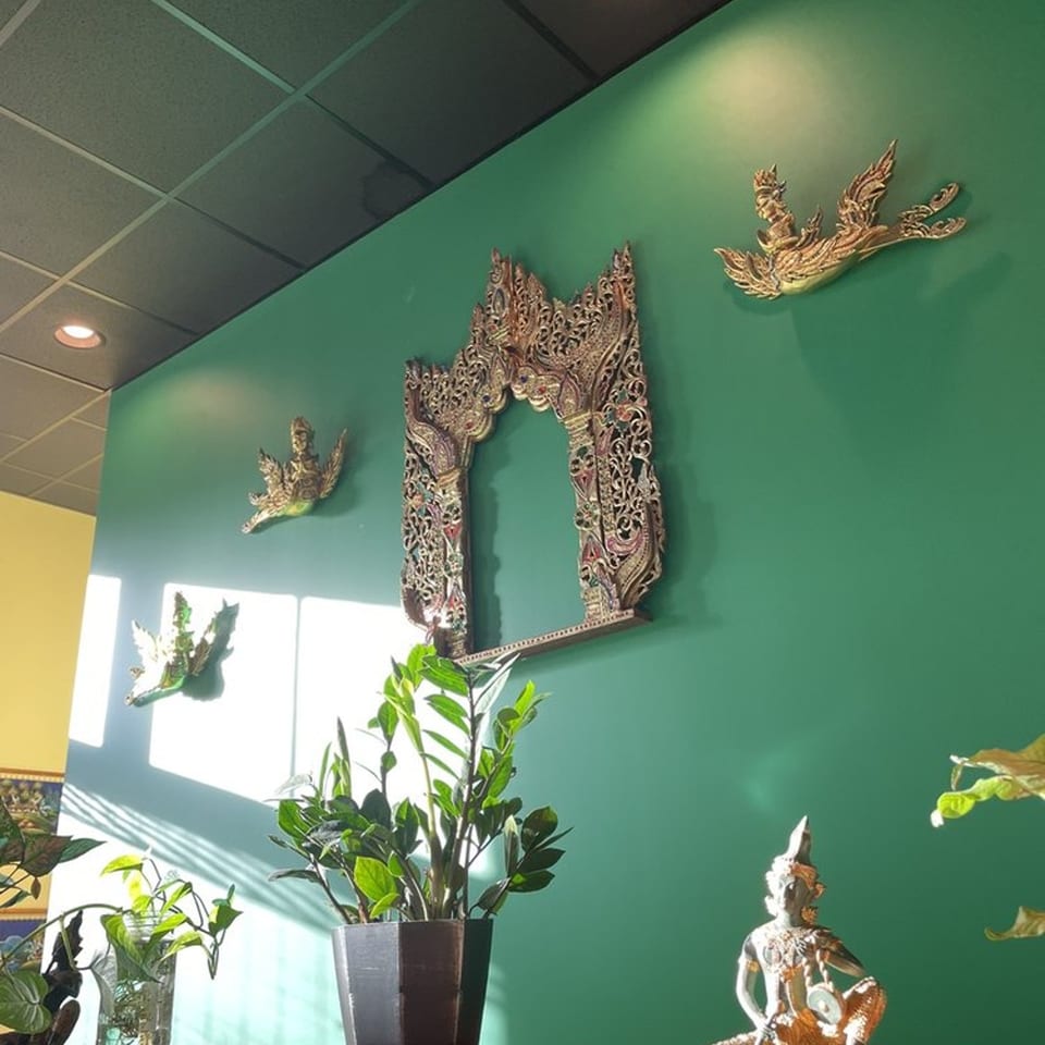 Green wall with ornate golden Thai decor and potted plants in a sunlit restaurant interior.