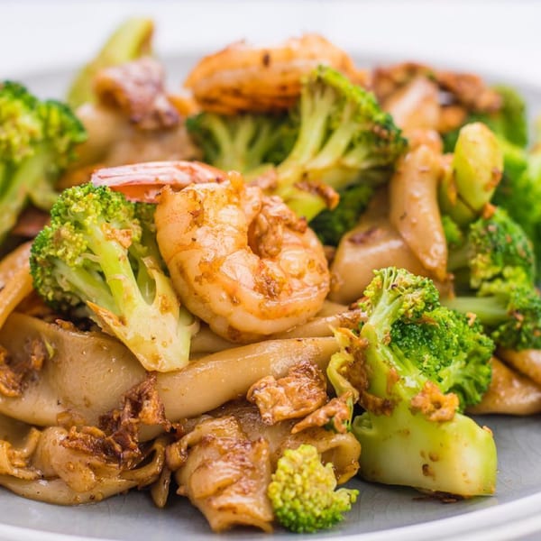 Close-up of a Thai dish with shrimp, broccoli, and flat noodles on a white plate.
