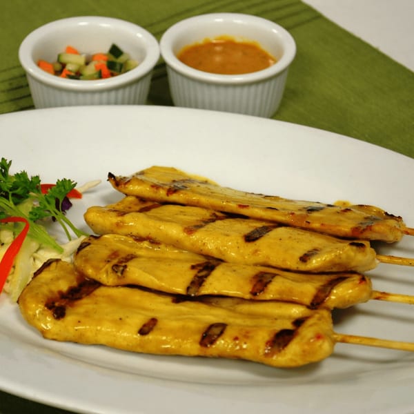 Grilled chicken satay skewers with peanut sauce and cucumber salad on a white plate.