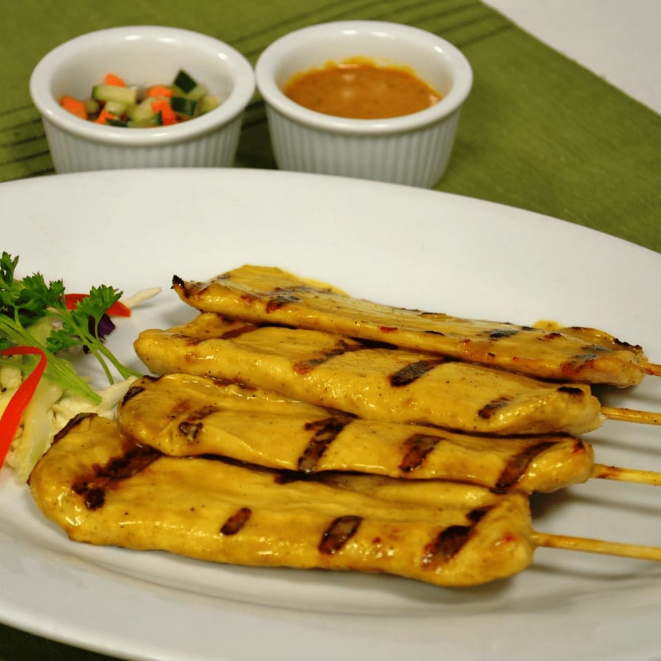 Grilled chicken satay skewers with peanut sauce and cucumber salad on a white plate.
